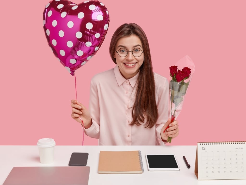 Mujer sonriente con regalo y rosas de San Valentín en oficina, simbolizando compromiso laboral.